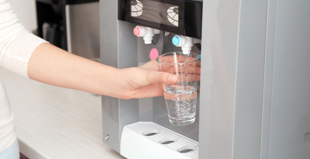 Gros plan sur des mains tenant un verre sous un distributeur d'eau, arrière-plan flouté d'un bureau lumineux